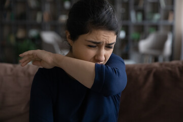 Vampire cough. Sick millennial indian woman staying ill at home sitting on sofa coughing covering...