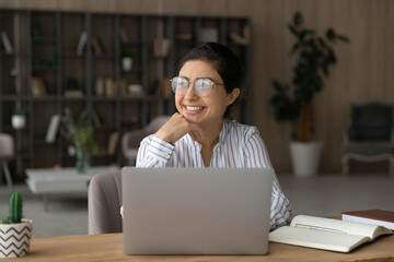 Young indian lady scientist in glasses work on pc do research sit by desk at home in library think...