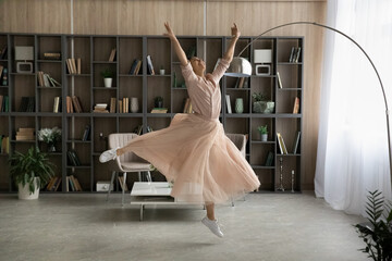 Embodied motion. Inspired teen indian lady in tender flowing dress comfy footwear dance ballet jump in air at large modern designed apartment. Happy young woman dancing alone at new home enjoy freedom - obrazy, fototapety, plakaty