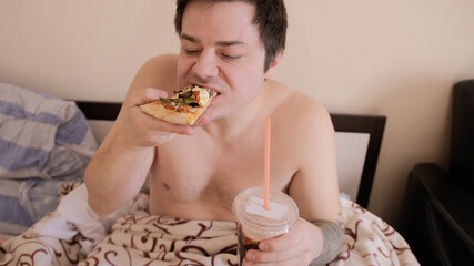 Breakfast in bed. The young man takes a bite of pizza and chews it, holding a milkshake in his other hand