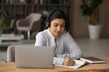 Young indian woman take part in studying online use headphones pc handwriting learning material...