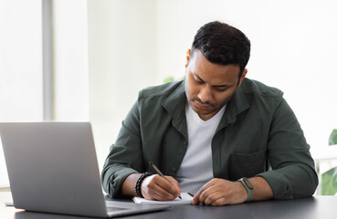 Concentrated Indian man taking notes while sitting at table with laptop at home or office, young...