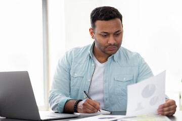 Pensive Indian young entrepreneur looking through paperwork while sitting at the desk at home or...