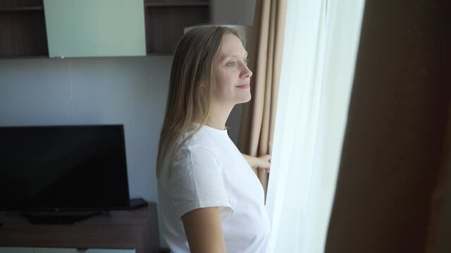 Happy optimistic young woman in white t-shirt, parting the beige curtains for the morning sun to illuminate her cozy apartment.She looks out the window alone and enjoys beautiful view of the cityscape