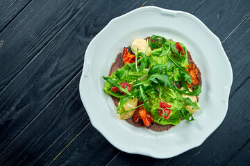 The traditional Italian appetizer salad is vitello tonnato. Thinly sliced beef with rucola, tomatoes and pesto, served in a white plate on a dark background