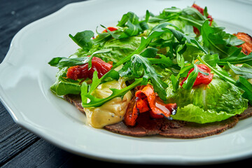 The traditional Italian appetizer salad is vitello tonnato. Thinly sliced beef with rucola, tomatoes and pesto, served in a white plate on a dark background