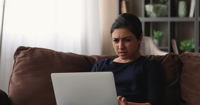 Indian woman sit on sofa work on laptop feels desperate having unexpected device malfunction. Female read awful online e-mail news. Unsaved important information, need pc repair, system error concept