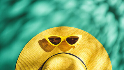 Yellow straw hat and sunglasses over green turquoise background with sunlight and tree shadow...