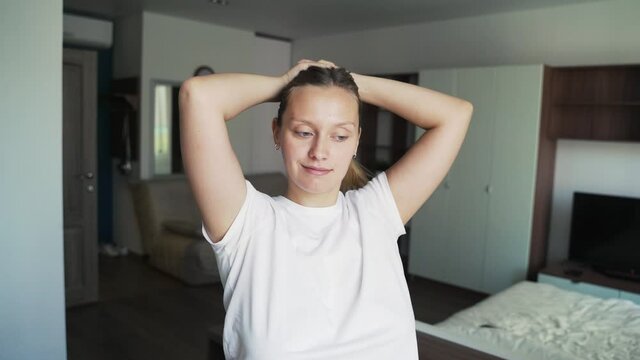 Happy beautiful blonde puts on an scrunchy on her long hair, makes a ponytail and smiles, standing in front of a window in a bright living room. Pulls smooth hair into a ponytail. Front view