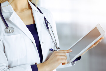 Unknown woman-doctor is holding a tablet computer in her hands, while standing in a sunny clinic cabinet. Female physician at work, close-up. Perfect medical service in a hospital. Medicine concept