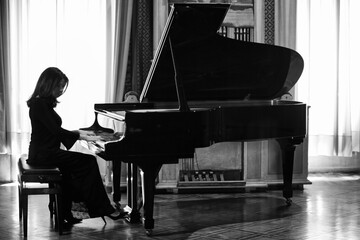 An elegant female pianist plays a grand piano. Black and white photo of a young musician in a long...