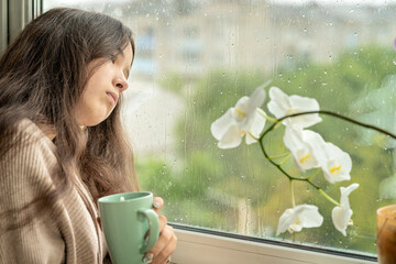 Girl drinking coffee looking at rain through a window at home or hotel. Girl with cup of coffee or tea near the rainy window look at the street.