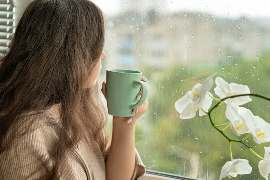 Girl drinking coffee looking at rain through a window at home or hotel. Girl with cup of coffee or tea near the rainy window look at the street.