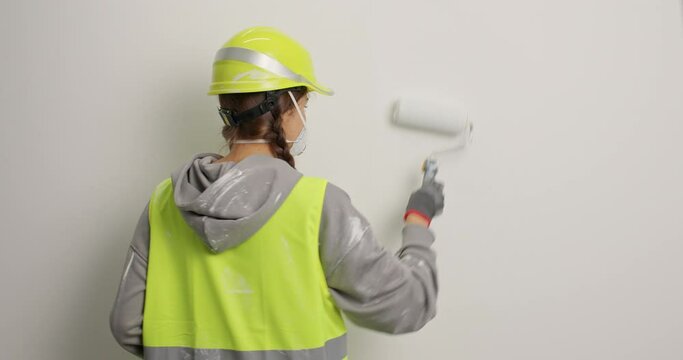 Room painting repair and design concept. Woman builder or decorator paints walls with roller wears hardhat and uniform turns gaze at camera asks what remodels freshy built house. Painter at work
