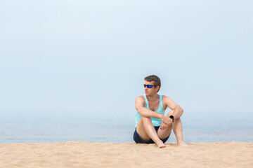 Young adult man sitting alone on sand and looking far away on side at calm sea beach in warm sunny morning. Thinking about life. Peaceful atmosphere in nature. Empty place for text, quote or sayings.