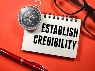 Education concept.Text ESTABLISH CREDIBILITY on notebook with compass,pen and glasses on red background.