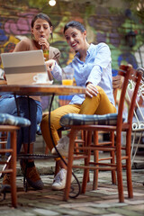 two caucasian young adult female friends sitting in open door cafe, looking something on laptop, smiling.