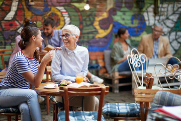 two females, different ages, sitting in outdoor cafe, talking, smiling, drinking coffee, looking each other.