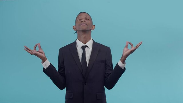 Relaxed black male businessman in a suit and with eyes closed holds hands a gesture from yoga namaste in the studio on blue background. Concept of calming and resting from work.