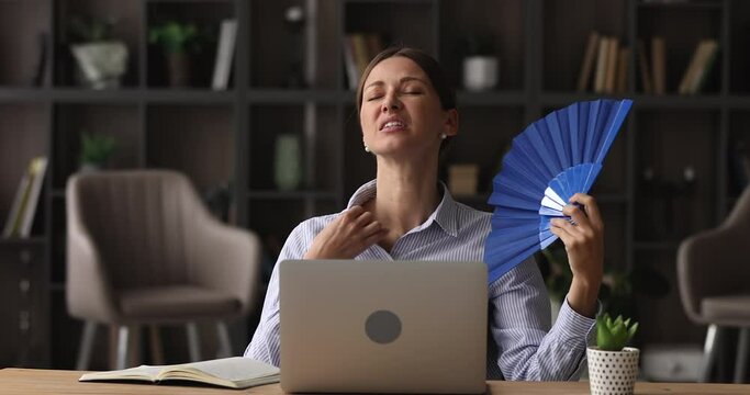 Woman sit at desk use blue fan cools herself, feels unwell, suffers from heat inside of office room without air-conditioning system. Excessive sweating, sensitivity to heat, hormonal imbalance concept