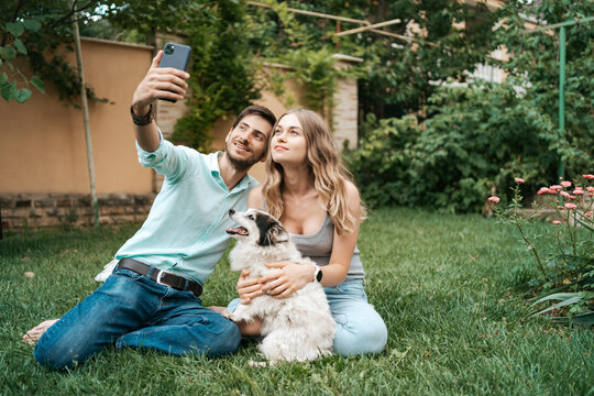 Beautiful happy couple making selfie with their lovely dog on the backyard while sitting on the grass