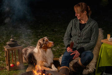 Young woman drinks red wine by the campfire in the forest. Her Australian Shepperd is lying next to her on a rug in the grass. The dog looks at its owner. Enjoying food and drink in leisure time in
