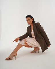 Elegant woman in peach color silk slip dress and brown squared wool blazer sitting and posing in...