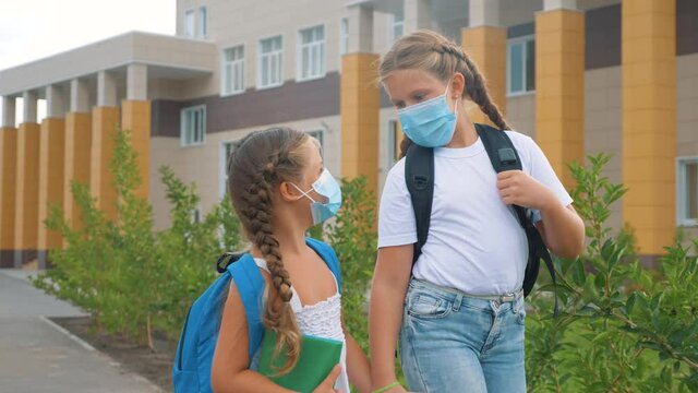 Schoolgirls with backpacks go through the schoolyard. Children at school in protective masks. New normalcy in schoolchildren. children kolniki go to school in medical masks, at a safe distance