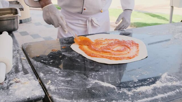 Pizzaiolo putting sauce on dough pizza on the table. Italian food in outdoor restaurant kitchen. Slow motion. Closeup of hands spreading tomato red sauce 