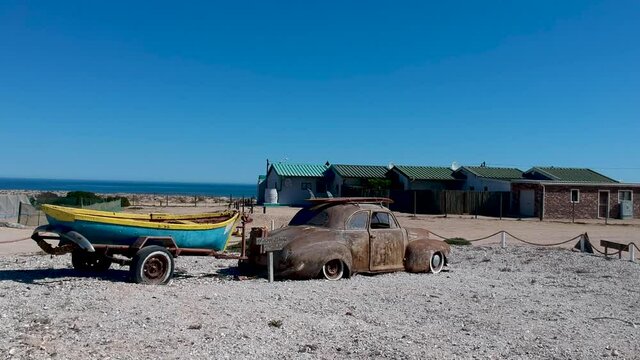 Rusty car with surfboards on the roof old trailer with a boat on sea shells by the sea buildings in background sign says topless photos only camera approaching from behind car aerial video.