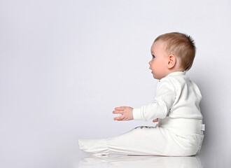 Small cute baby boy in light cotton overalls. Jumpsuit sitting on the floor and looks away over...