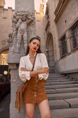 Elegant woman in white blouse and brown skirt standing leaning on grey stone fence at stairs...