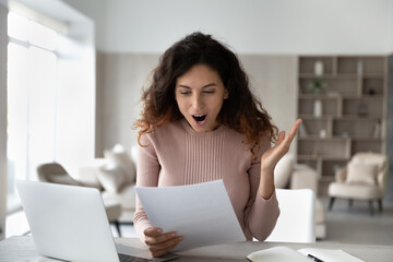 Happy excited Latino woman shocked by good news in paper correspondence working online on laptop at...