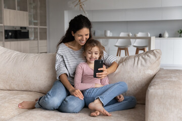 Obraz premium Smiling young Hispanic mother and teen daughter relax on sofa at home talk speak on video call on cellphone. Happy Latino mom and little girl child have fun use smartphone have webcam online event.