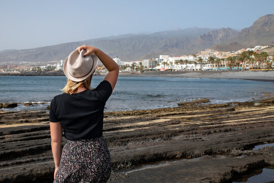 Rear view of a young woman holding her straw hat with her hand and looking at the entire resort of Costa Adeje from the rocky eroded coast of Playa de Las Americas, in Tenerife, Canary Islands, Spain