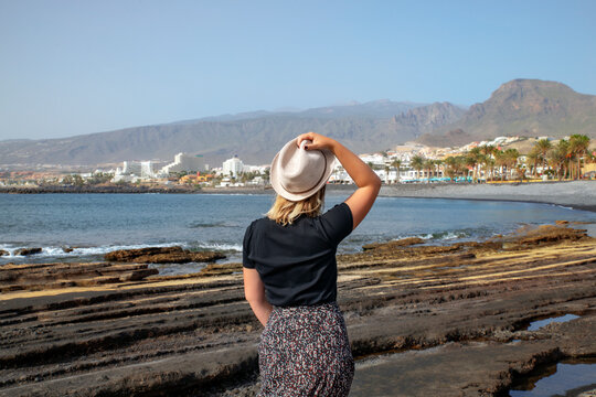 Rear view of a young woman holding her hat with one hand and looking at the entire resort of Costa Adeje from the eroded rocks on the coast of Playa de Las Americas in Tenerife, Canary Islands, Spain