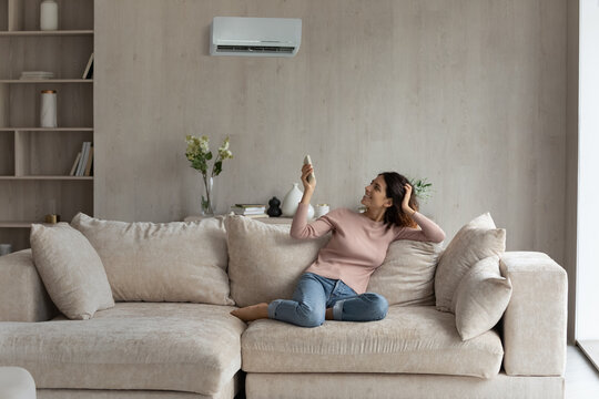 Young Latino Woman Relax On Sofa In Living Room Turn Electronic Conditioner With Remote Breathe Fresh Ventilated Air. Millennial Hispanic Female Renter Tenant Use Condition Device At Home Resting.