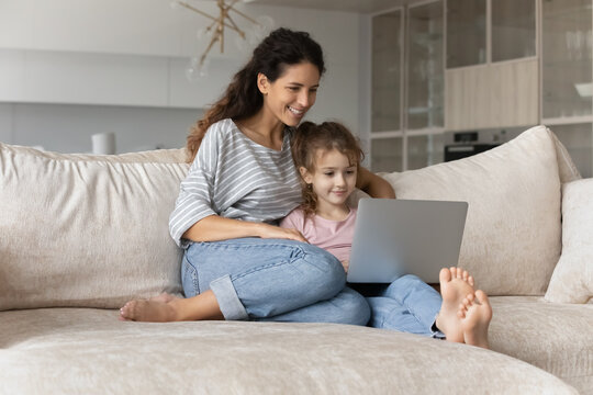 Happy Young Hispanic Mother And Small Biracial Daughter Relax On Sofa In Living Room Using Computer Together. Smiling Latino Mom And Ethnic Girl Child Rest On Couch Talk On Video Call On Laptop.