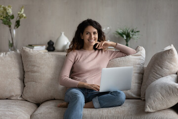 Portrait of smiling young Latino woman sit on sofa at home use modern computer gadget work online. Happy millennial Hispanic female relax on couch in living room with laptop. Technology concept. - Powered by Adobe