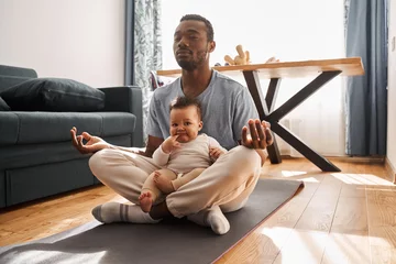 Abwaschbare Fototapete Man in casual clothes meditating with his son and sitting on lotus pose at home © Yakobchuk Olena
