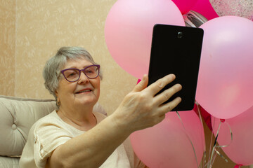 A cute senior lady holds a bunch of balloons in one hand and a tablet in the other to wish her...