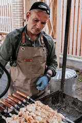 BBQ party in the backyard. A man in a baseball cap and apron prepares a chicken shish kebab for a...