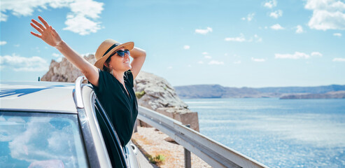 Cheerful Woman portrait enjoying the seaside road trip. Dressed a black dress, straw hat and sungllasses she wide opened arms and shining with happiness. Summer vacation traveling by auto concept.