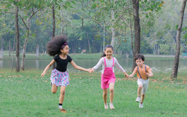 Naklejka na ściany i meble Adorable children hold hand together enjoy running in green park on holiday, group of primary school friendship play in garden, children Afro mix race girl and Asian kids spent time with nature