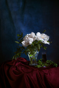 Dramatic photo in chiaroscuro style with white flowers of a tree peony on a dark background. still life with a glass of red wine and burgundy velvet drapery. Melancholy still life.selective focus.
