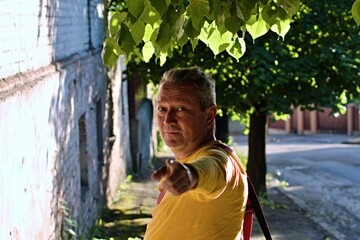 Man in yellow t-shirt in shade of trees stretched out his hand forward on blurred background of old city