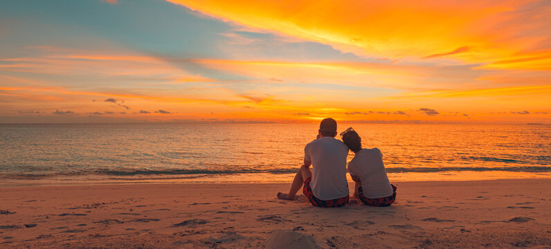 Romantic couple on the beach at colorful sunset on background. Beautiful tropical sunset scenery, romance couple sitting and watching the sunset sea and sky, horizon. Travel, honeymoon destination