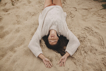 Beautiful woman lying on sandy beach, carefree tranquil moment. View above. Stylish young female in knitted sweater enjoying vacation and relaxing on coast