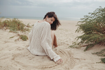 Beautiful woman with windy hair sitting on sandy beach with green grass, carefree tranquil moment....
