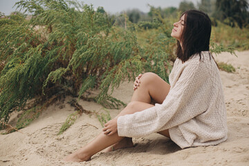 Beautiful woman with windy hair sitting on sandy beach with green grass, carefree tranquil moment....